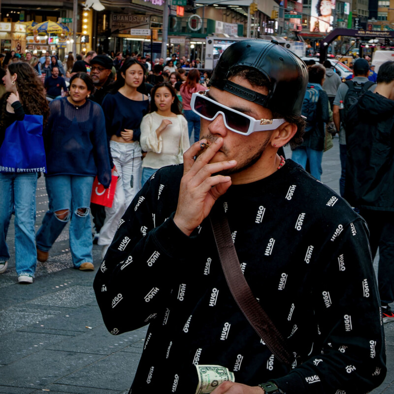 A man wearing a black HUGO sweater, white sunglasses, and a backward cap smokes a cigarette and holds cash in a busy city street, surrounded by pedestrians.