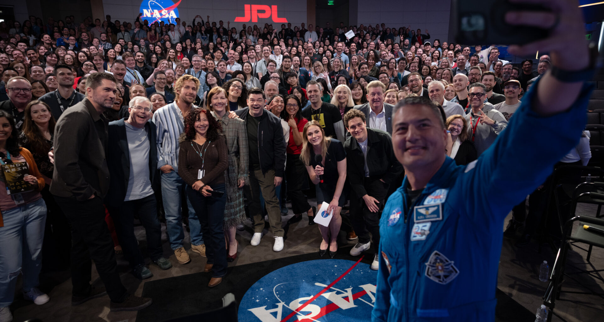 At right, a man in a blue flight suit with patches on it takes a selfie with his cell phone. He is in the photo, as well as panel members and the large audience. They are in an auditorium. In the far background, there is a NASA "meatball" insignia and the letters "JPL" on the wall.