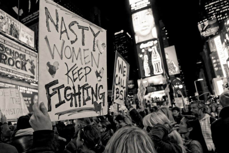 A crowd of protesters at night holds signs, including one that reads “Nasty women keep fighting,” in a brightly lit city area filled with billboards. The atmosphere is energetic and determined.