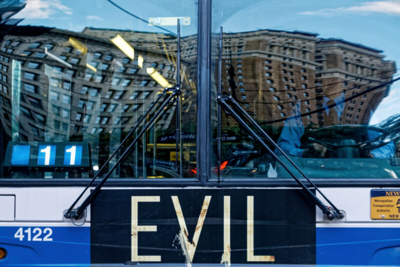 Close-up of a bus front with the word "EVIL" displayed on its sign, reflecting distorted images of tall city buildings in the windshield. The bus is marked with the route number 11 and ID number 4122.