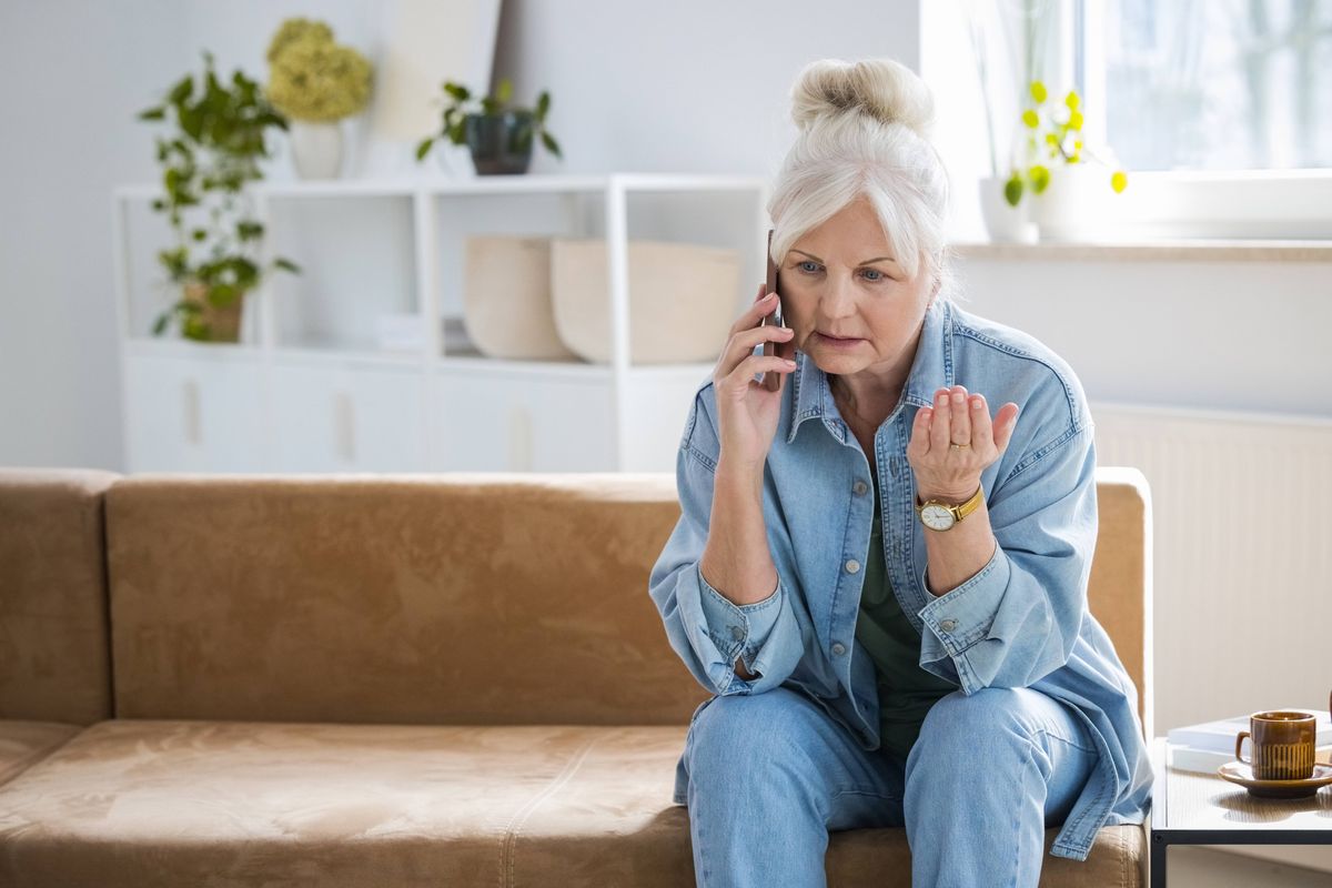 Angry senior woman wearing denim shirt sitting on sofa at home and using mobile phone.