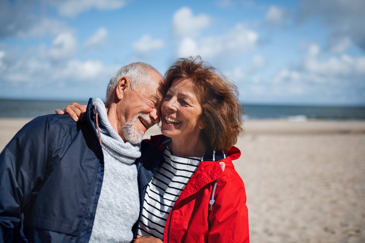 An elderly couple stands together on a beach, sharing a moment of happiness. The man is dressed in a dark jacket and scarf, while the woman wears a striped shirt and a red jacket. The background features a serene sea and a partly cloudy sky, suggesting a peaceful and joyful outdoor setting.
