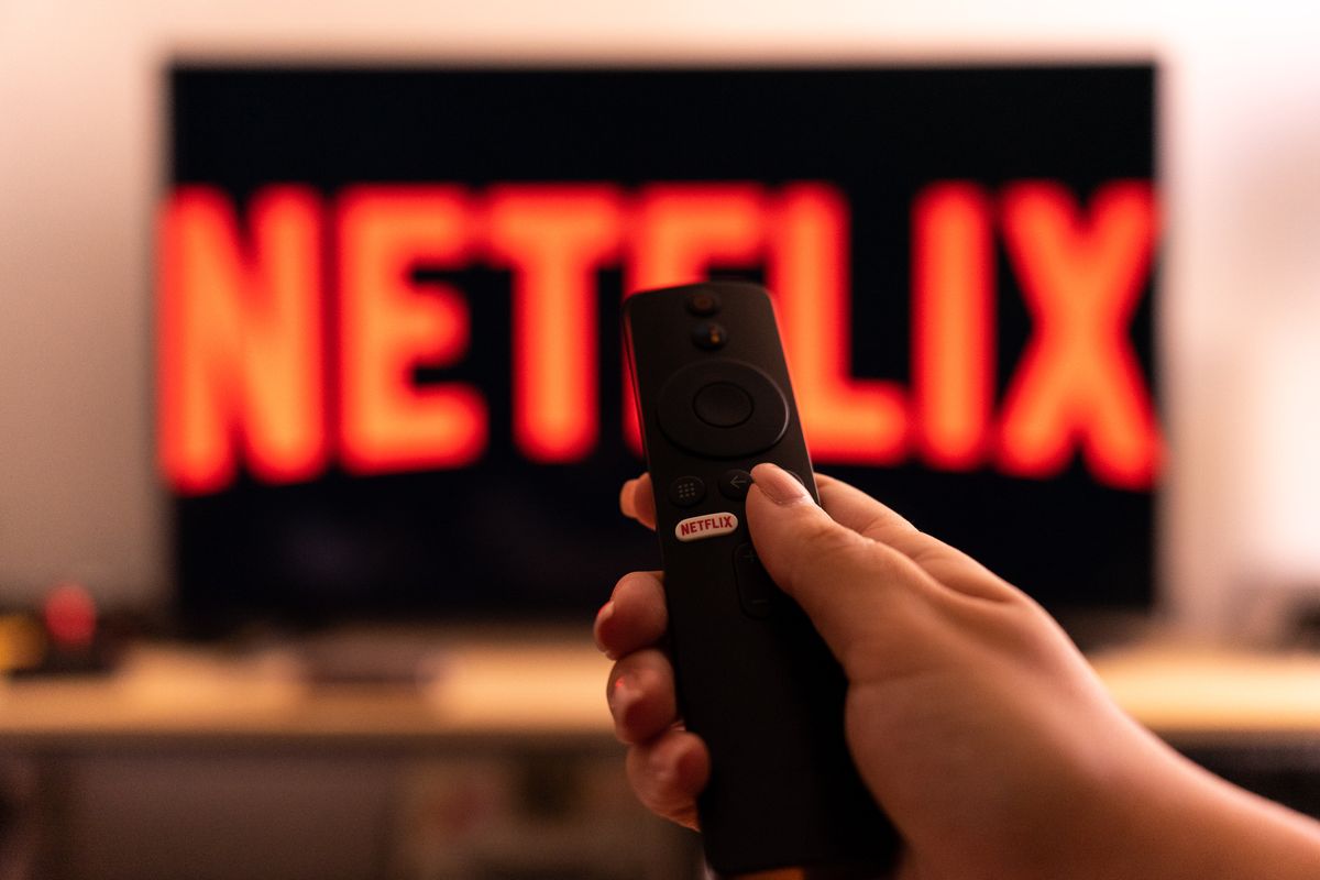 A hand holding a tv remote with a ''Netflix button'' is seen in front of a tv screen with the logo of Netflix. 