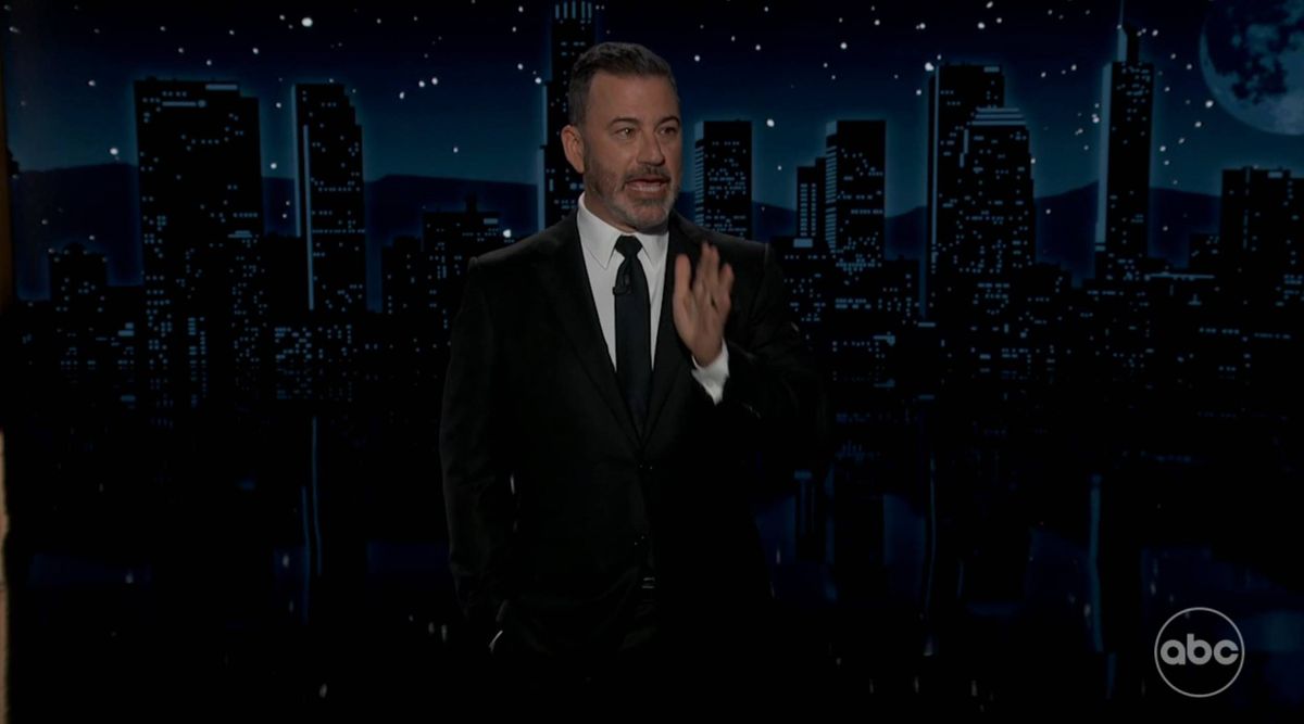 An individual dressed in formal attire, including a suit and tie, is standing in a dimly lit environment with an urban skyline visible in the background. The individual appears to be engaged in a speech or presentation.