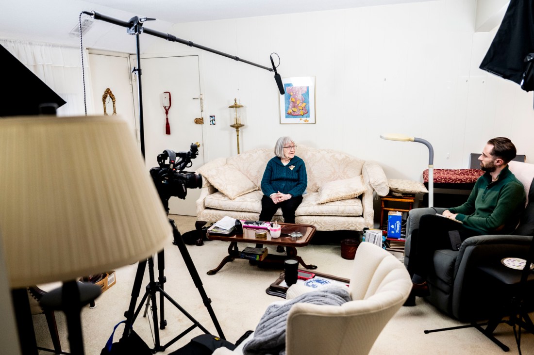 Ruth Gove seated on a white sofa in her home. She has a microphone above her. Cody Mello-Klein, the interviewer, sits in an armchair to the side.