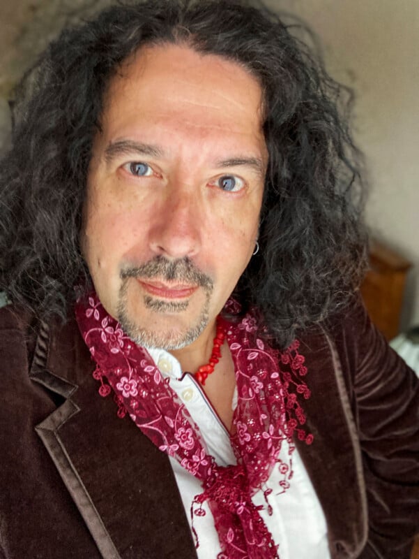 A person with curly dark hair and a goatee wears a brown velvet jacket, white shirt, red beaded necklace, and a sheer red scarf with floral embroidery, looking directly at the camera.