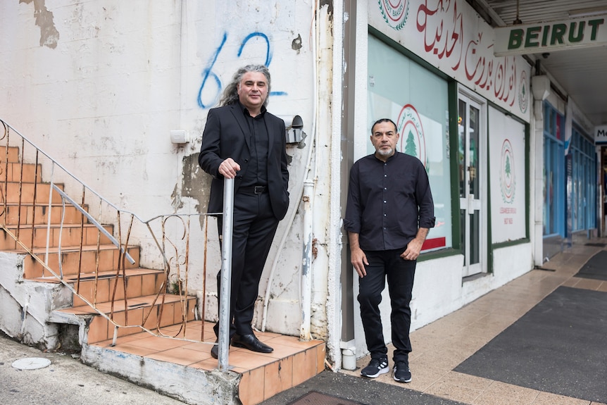 Two men dressed in dark blue suits stand on corner of gritty street near stairs and shop-front.