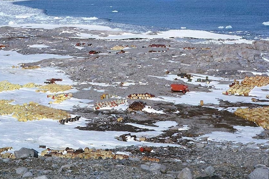 Fuel drums and other debris scattered in a snowy and rocky landscape near a coastline.