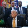 President Trump sits at his desk in the Oval Office of the White House on January 14. Wearing a navy blue suit and gold tie, he is surrounded by many people standing behind the desk.