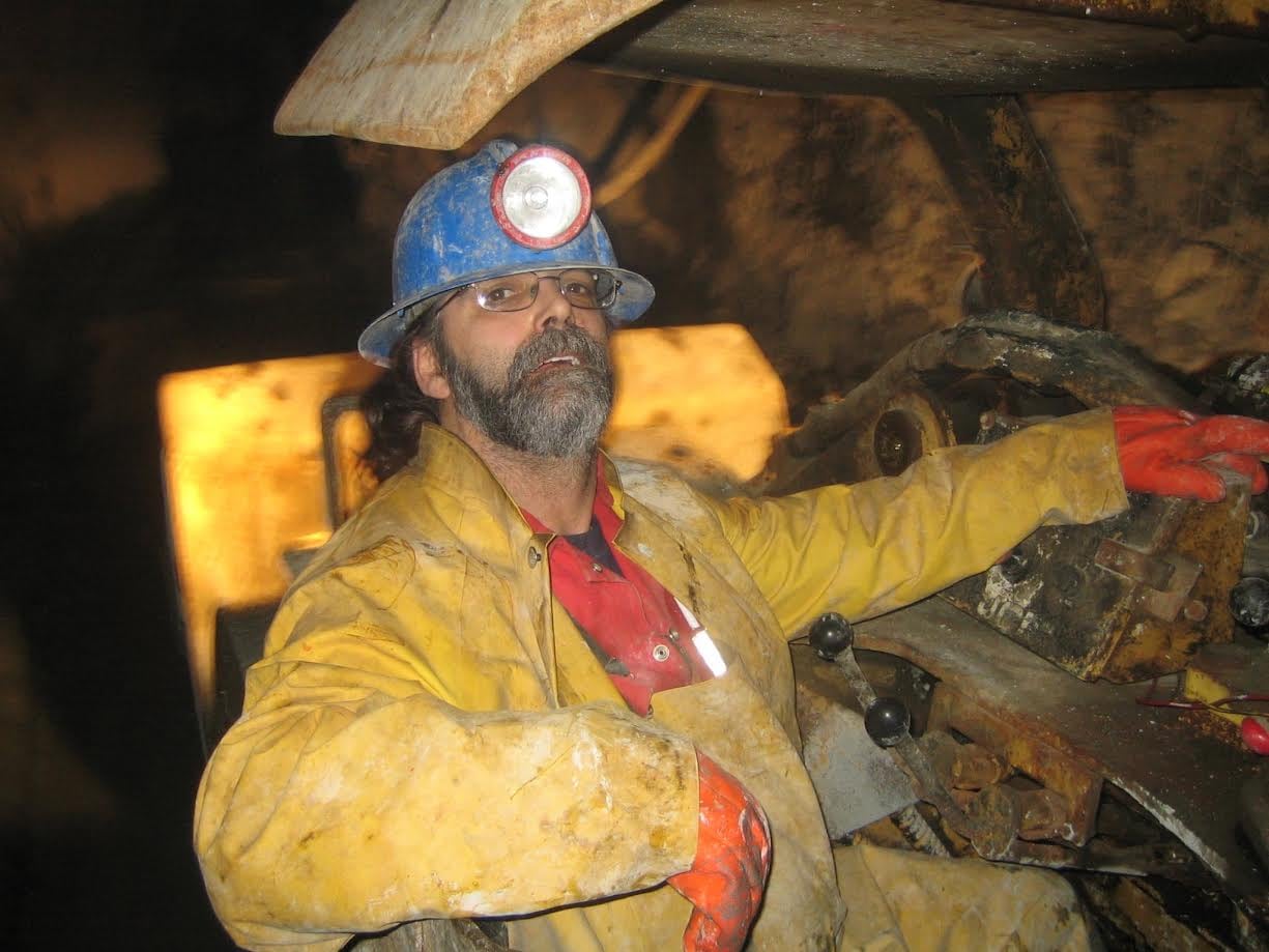 A bearded man wearing a hard hat operates heavy equipment in a mine.