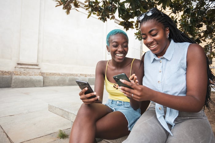 Two friends smiling and laughing while reading something on their smartphones.