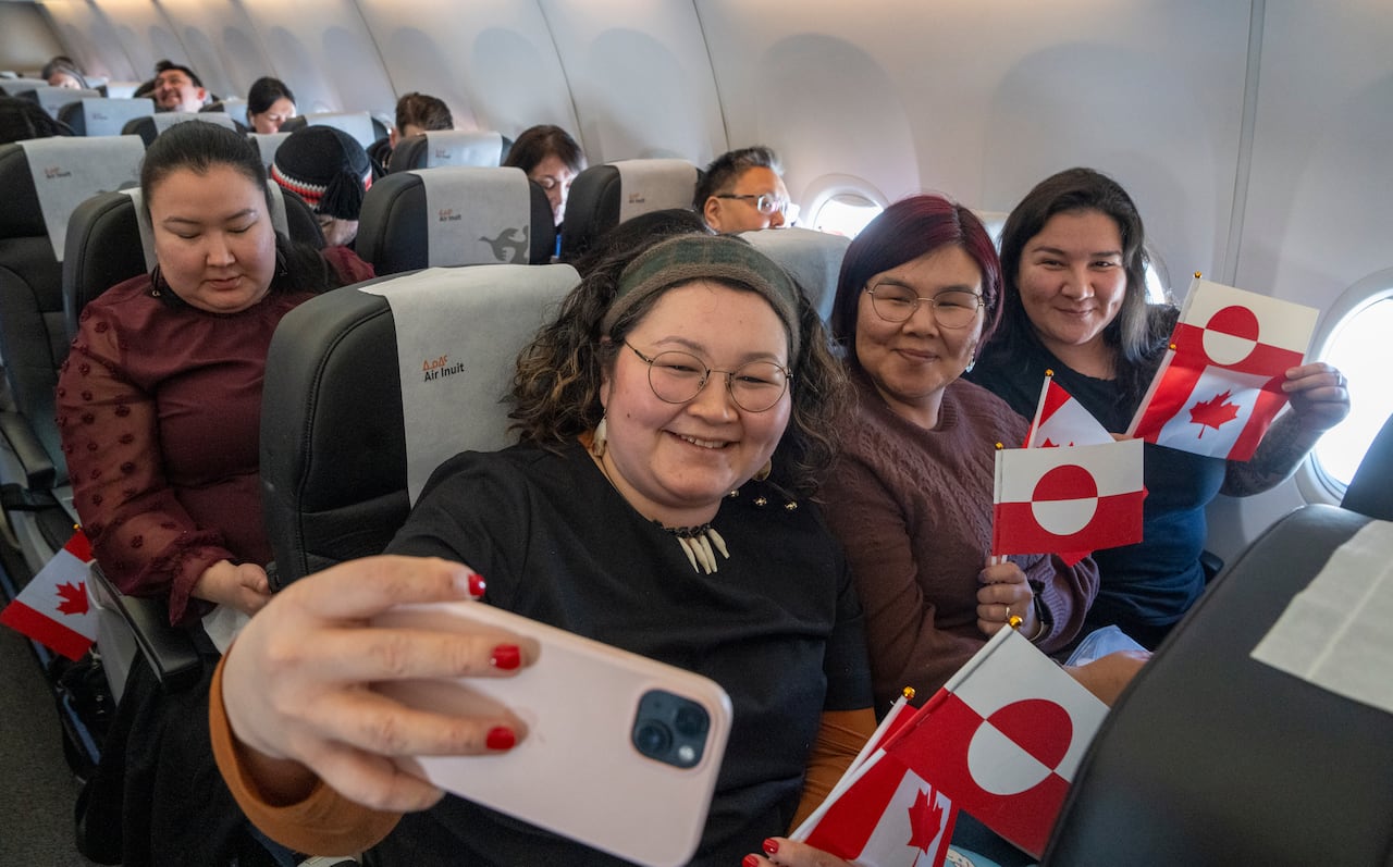 A group of women take a selfie on a plane while holding small Canada and Greenland flags.