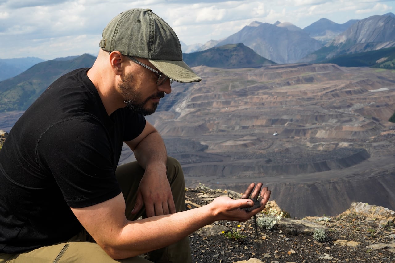 A bearded man in glasses wearing a cap and a black t-shirt holds a piece of coal with a terraced open-pit mining site in the background.