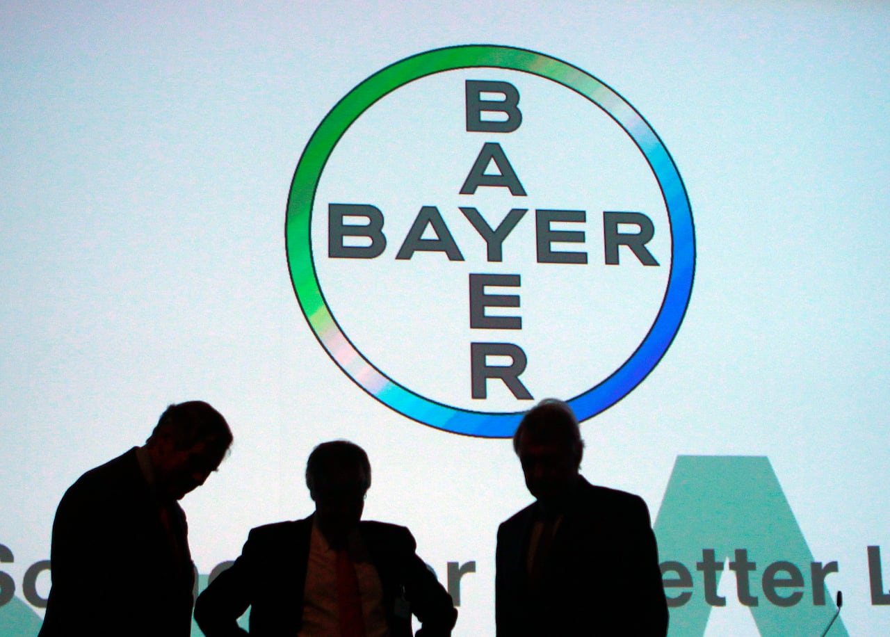 The silhouettes of three businessmen stand beneath a large logo for the pharmaceutical company Bayer AG.