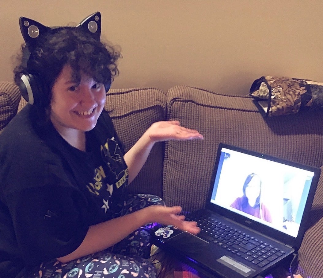 A curly-haired person wearing cat ear headphones gestures to a computer screen showing another person on camera.