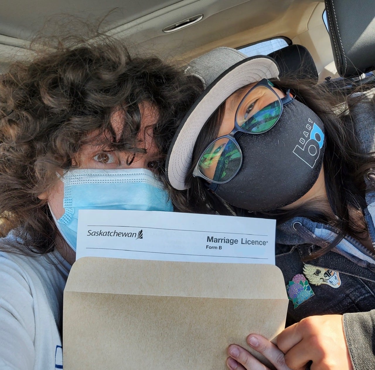 Two people in masks hold a brown envelope with a marriage licence document peeking out.