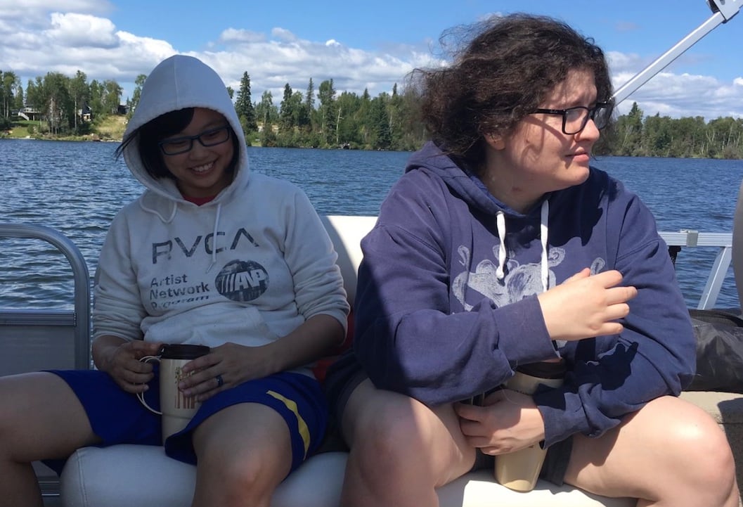 Two people sit beside each other on a boat on a sunny day, with a forested shoreline behind them.