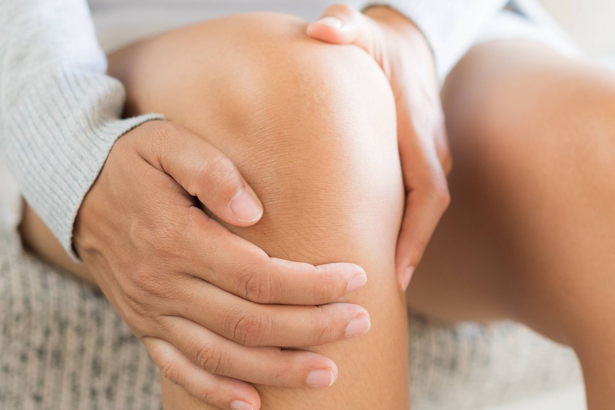 Closeup young woman sitting on sofa and feeling knee pain and she massage her knee at home. Healthcare and medical concept.