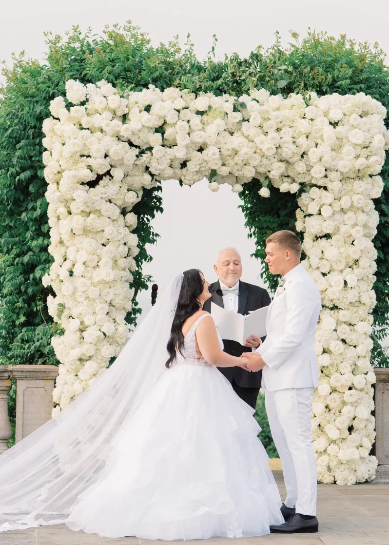 A couple exchanging vows in front of a floral arch with an officiant at their wedding. The bride wears a flowing gown; the groom is in a white suit