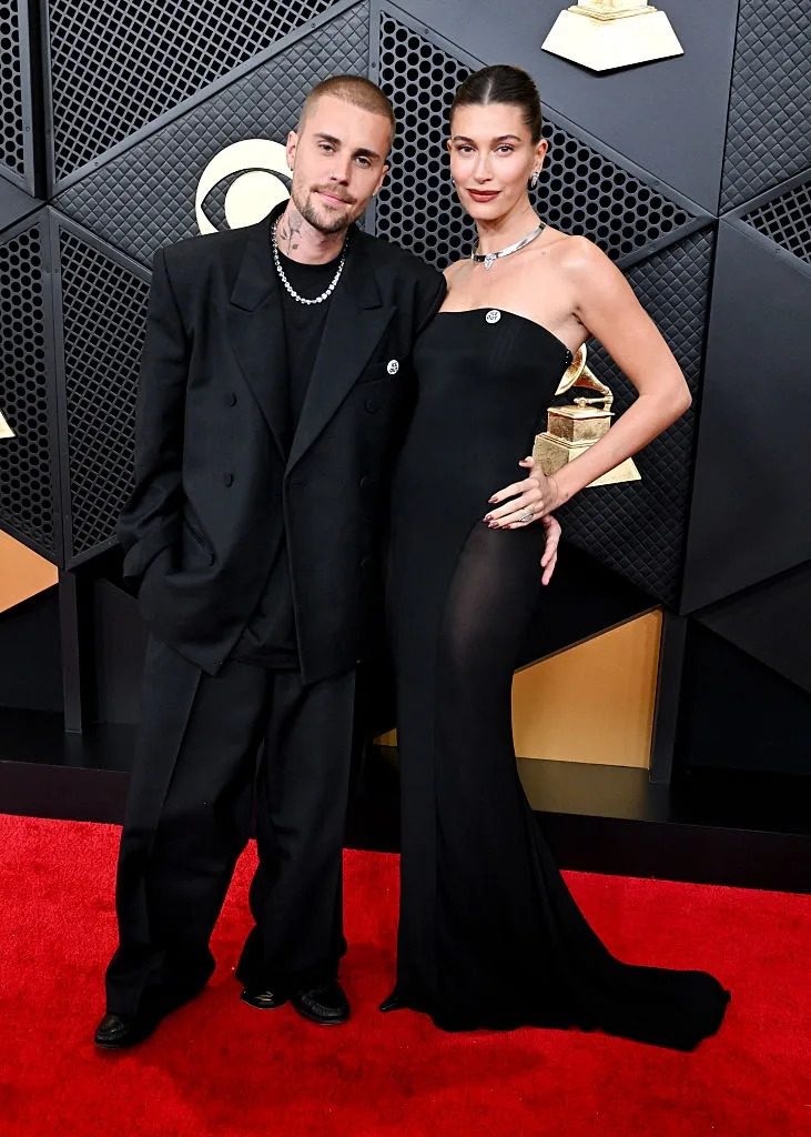 Two people on a red carpet; one in a loose black suit, the other in a sleek black strapless gown, both wearing necklaces