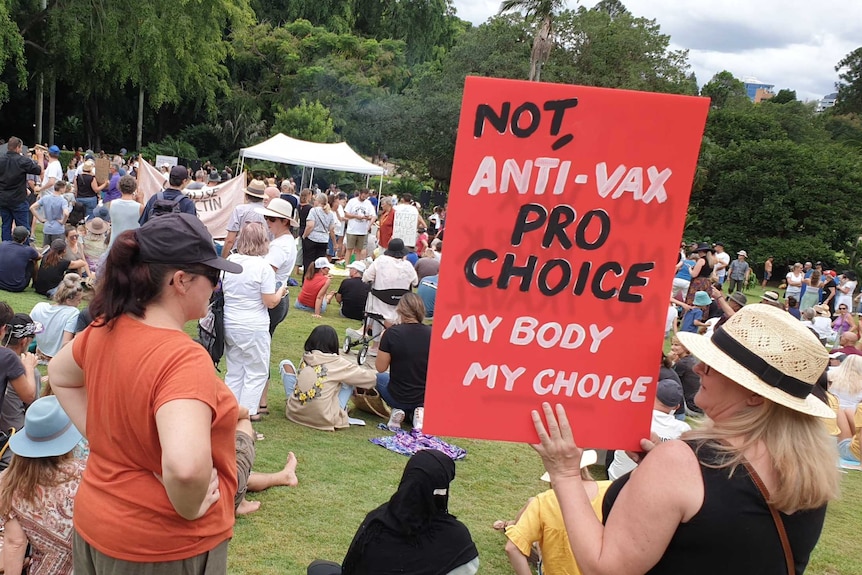 Protesters gather in a park, with one woman holding a red sign reading "Not anti-vax, pro choice, my body my choice"