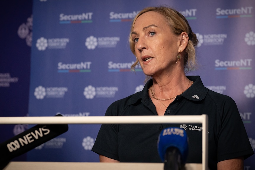 A woman with blonde hair, speaking into news microphones at a press conference.