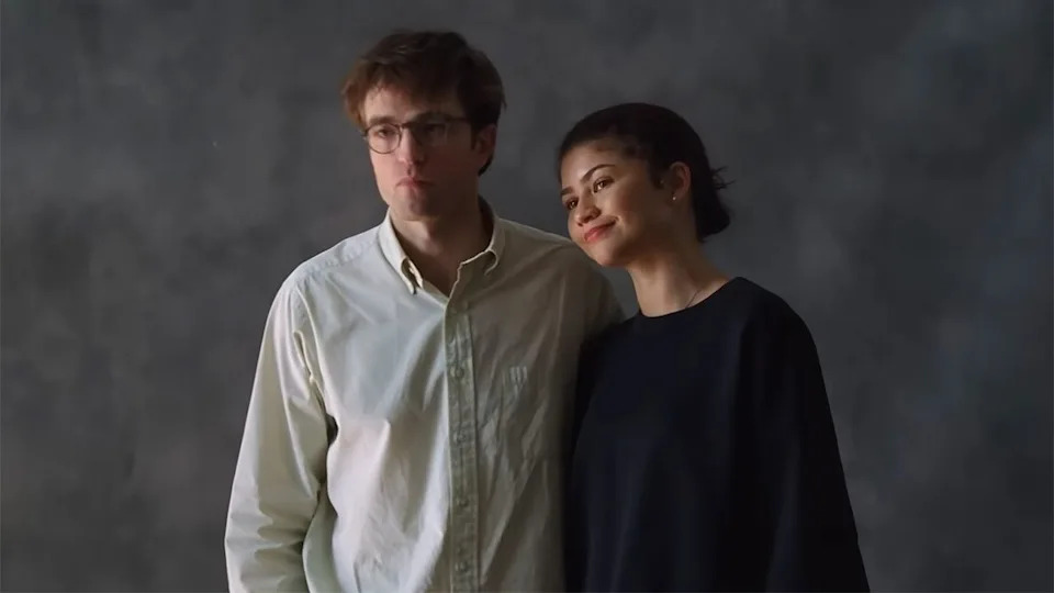  Robert Pattinson in a white button down and Zendaya in a black sweatshirt standing in front of a plain grey background. . 