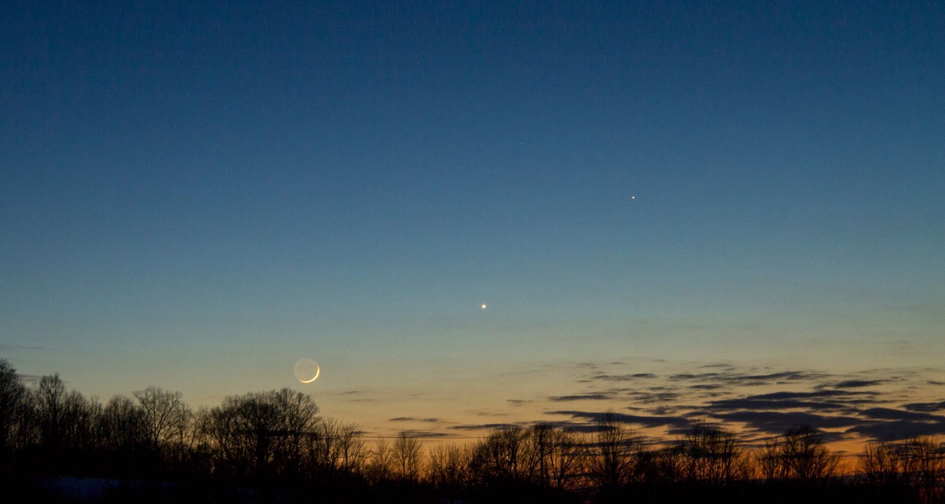 A orange and blue night sky with a glowing crescent moon and a bright dot for the planet Mercury with a series of silhouettes of trees in the foreground