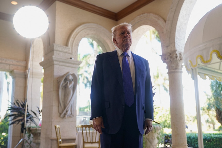 Donald Trump, wearing a blue suit, stands in front of archways