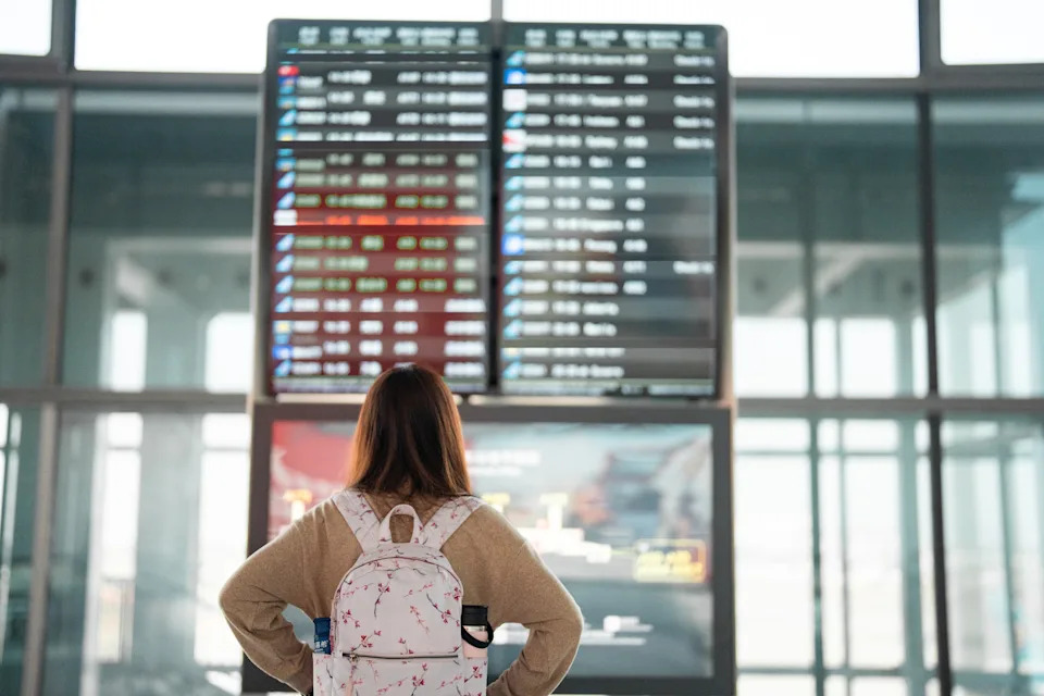 A person with a floral backpack stands in an airport terminal, looking at a large departure board with various flight information