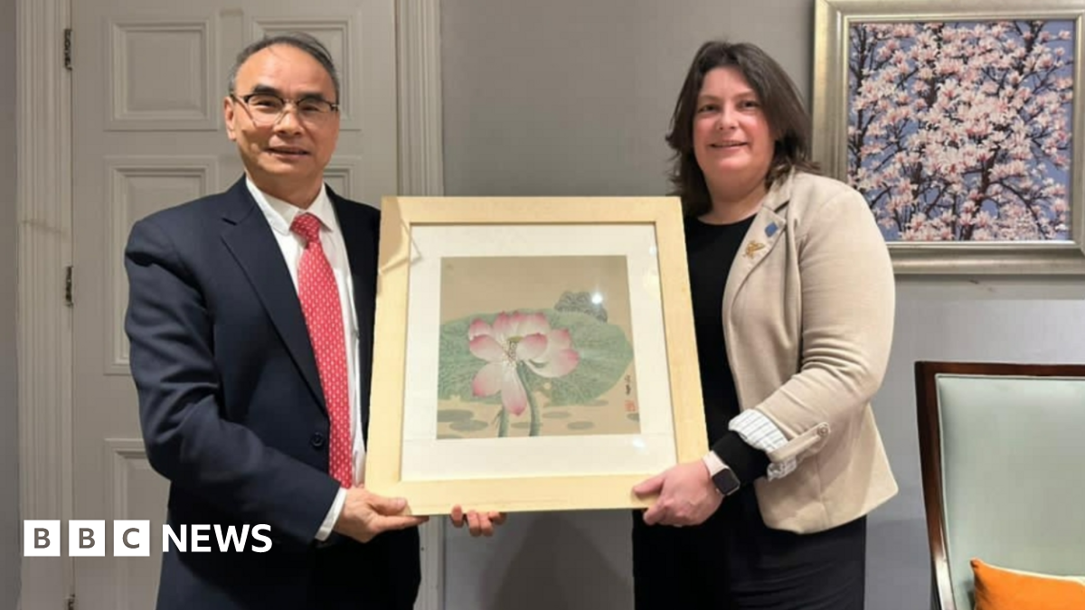 Hu Muqing, wearing a dark suit and red tie, and Laura Pye, wearing a black dress and cream jacket, hold a painting of a white and pink flower in a gold coloured frame during their meeting in China