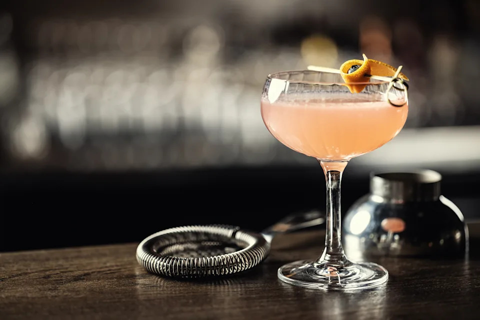 Cocktail glass with an orange twist garnish on a bar counter next to a metal strainer and shaker