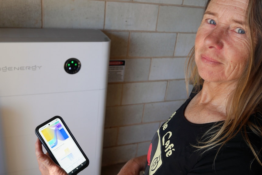 Picture shows a woman standing in front of a battery unit, she is holding a mobile phone in her hand that displays a colourful g