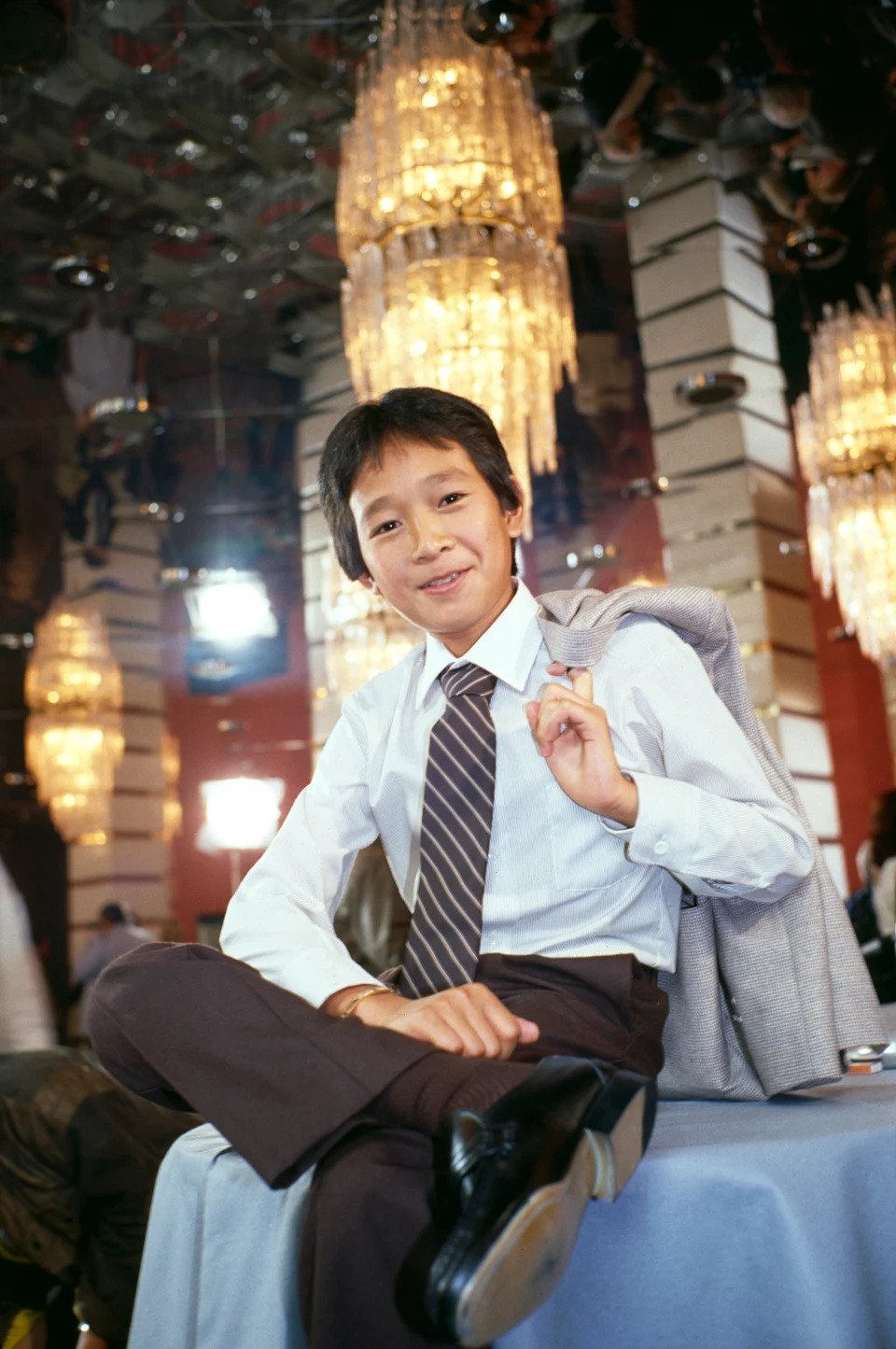 Person in a suit with a jacket over their shoulder, smiling and sitting with legs crossed in a room with chandeliers