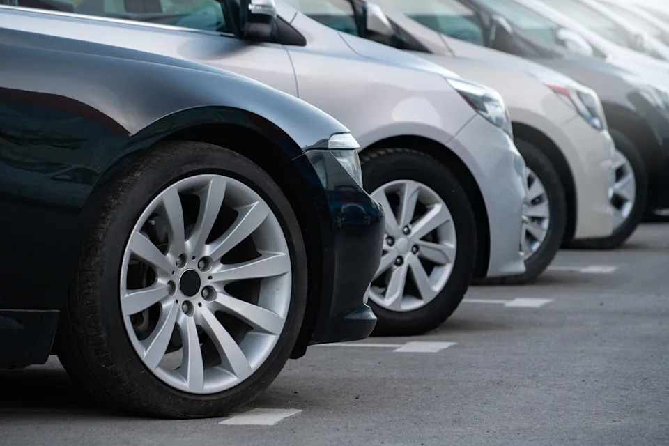 Several parked cars in a row in a parking lot, showcasing the side profiles and wheels of various models