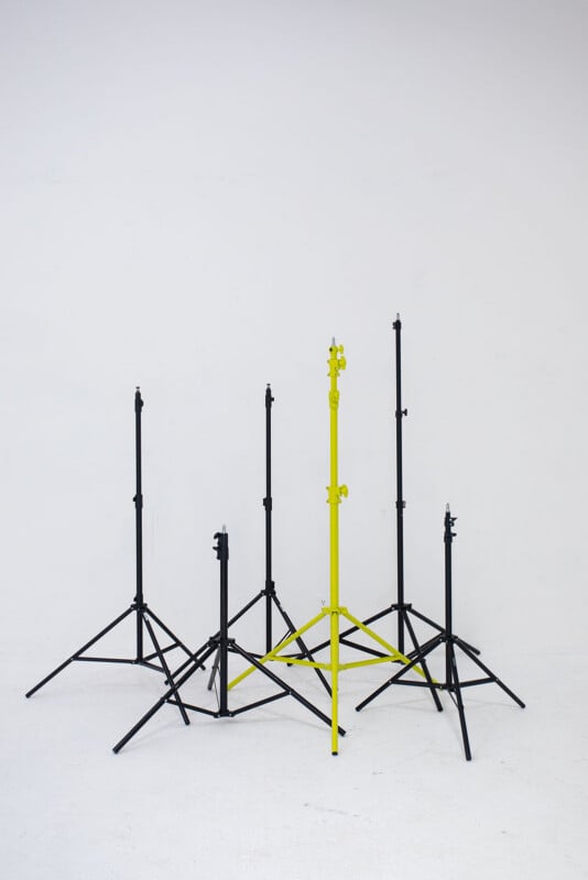 Six photography light stands on a white floor against a white wall. Five stands are black, and one is bright yellow, standing prominently in the center among the others.
