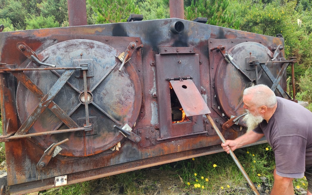 A large metal kiln with two drums. Matt bends down to open its door ready to feed the flames within