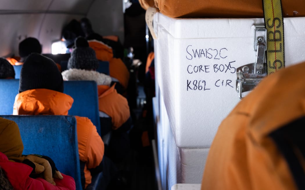 Interior shot of a small plane, showing the backs of passengers dressed in Antarctic field gear, with a white polystyrene box in the foreground labelled 'SWAIS2C CORE BOX 5'