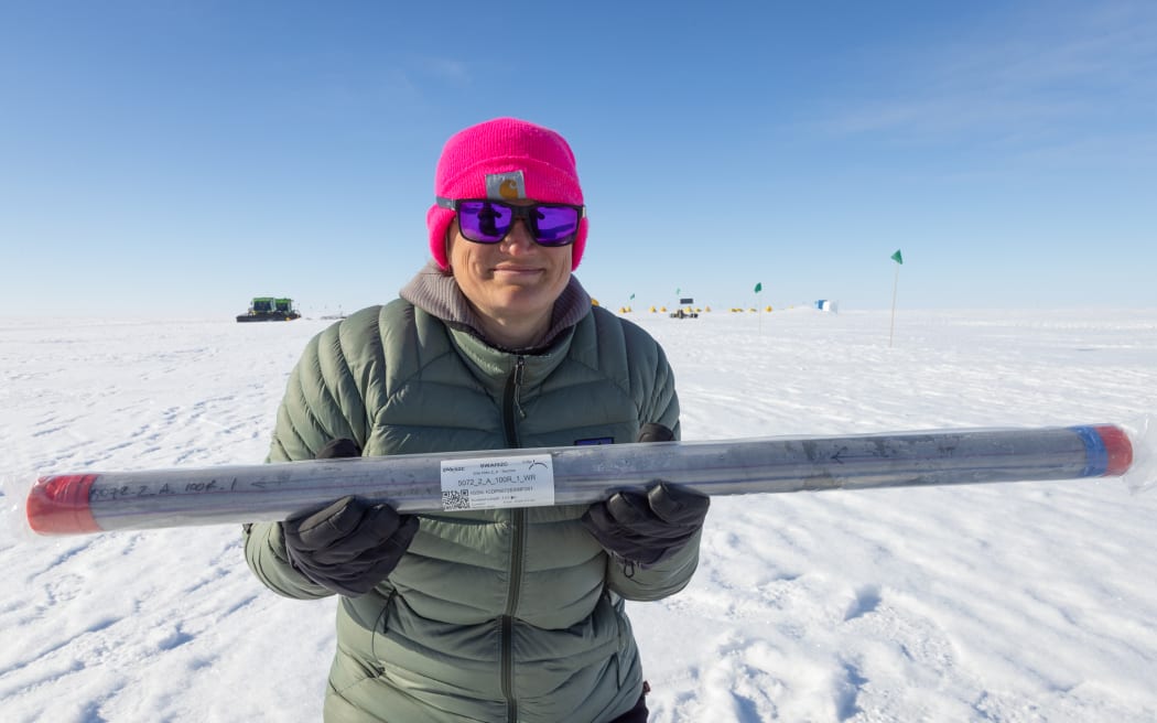 Molly Patterson, wearing field gear and a bright pink hat and standing on a snowy surface, smiles while holding a cylindrical tube containing a sediment core sample.