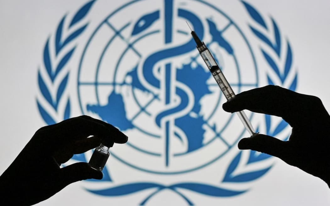 An illustrative image of a person holding a medical syringe and a Covid-19 vaccine vial in front of the World Health Organization logo displayed on a screen.