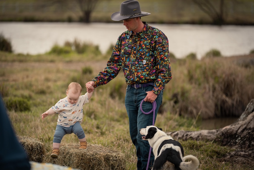 In one hand, Sam holds his baby's hand while he holds his dog on a leash in the other.