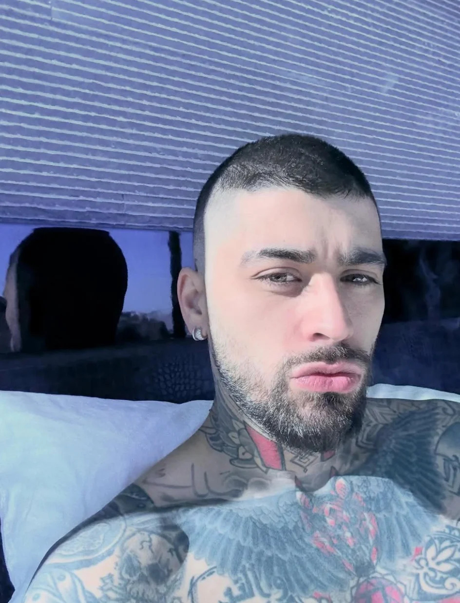 Person with tattoos is taking a selfie while lying on a bed, looking directly at the camera