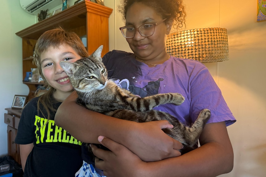 A young girl hold a relaxed cat, next to a smiling young boy. 