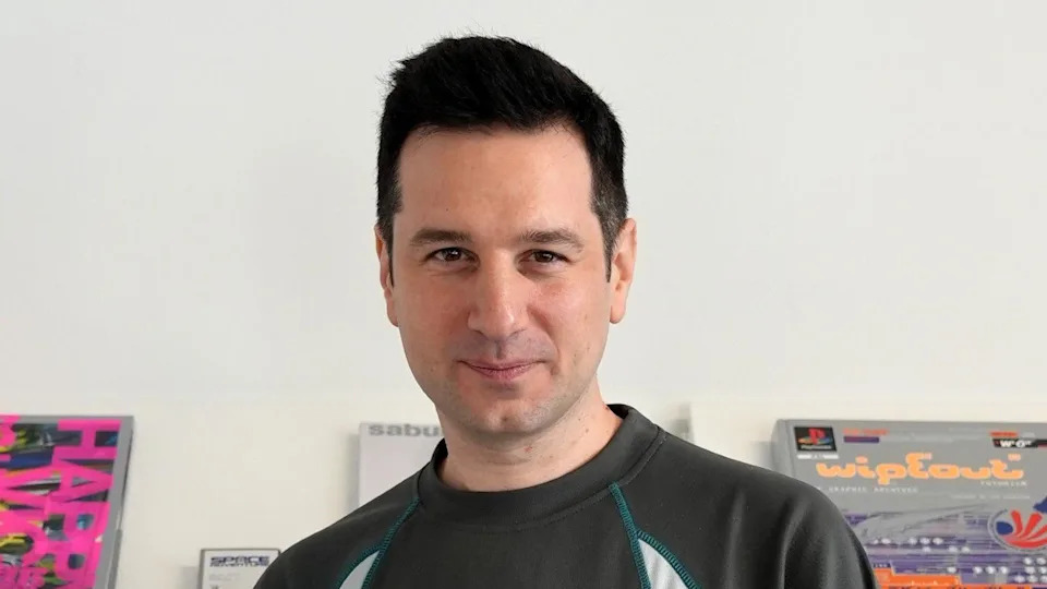 A man, Alexis Garavaryan is smiling and looking at the camera. In the background are books about gaming including the game Wipeout.