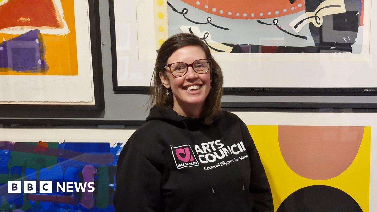 Suzanne, a woman with short brown hair, she wears glasses and has a bright smile as she stands in front of bright contemporary art.