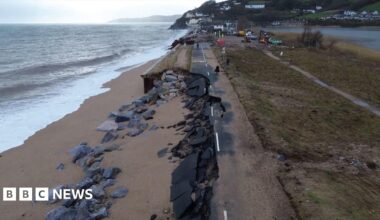 A picture of the washed away road. There is a number of rocks and sand covering the road next to the sea front.