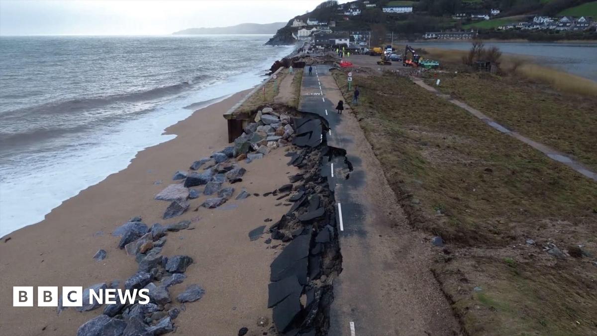 A picture of the washed away road. There is a number of rocks and sand covering the road next to the sea front.