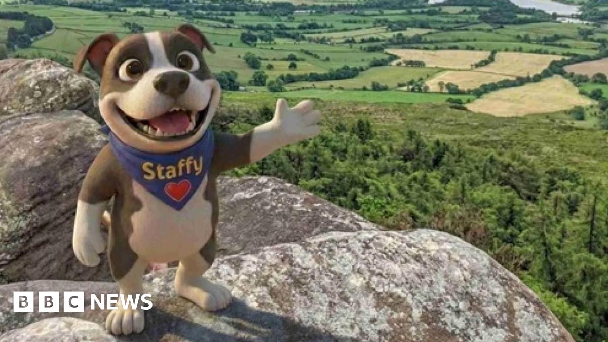 An animated design of a dog on its hind feet, waving with its paws towards the camera with a smile. The dog is brown and white with a blue neckerchief with "Staffy" and a heart on it. The dog stands on a rocky outcrop, overlooking green trees.