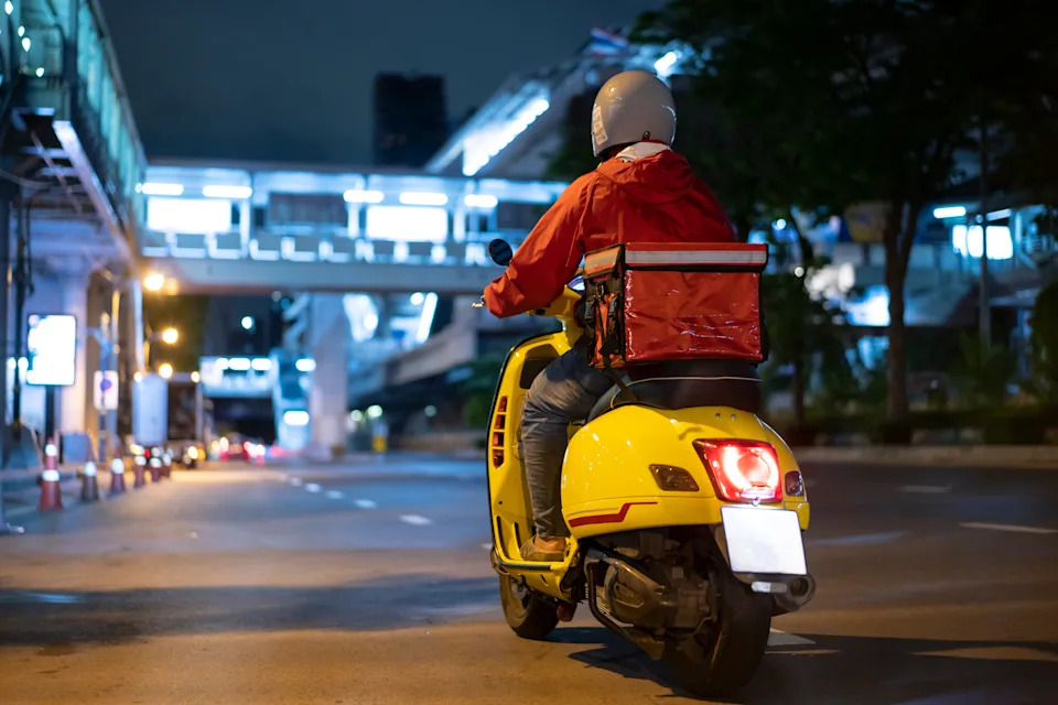 Delivery person on a yellow scooter rides through an urban area at night, focused on work and navigating city streets