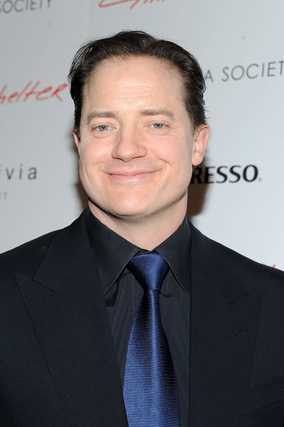Person in a formal suit and tie at an event, standing against a backdrop with text